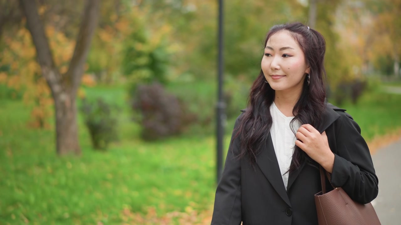 秋の小道に立つ、長い黒髪の若い女性のクローズアップ。黒いブレザーと白いシャツを着用し、ラペルに手を添えている。肩には茶色のハンドバッグを提げ、視線を向けている。