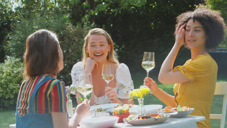 three female friends sitting outdoors in summer garden at home drinking wine and eating meal