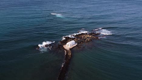 Tiny-white-chapel-surrounded-by-Mediterranean-sea-in-Crete,-aerial-view