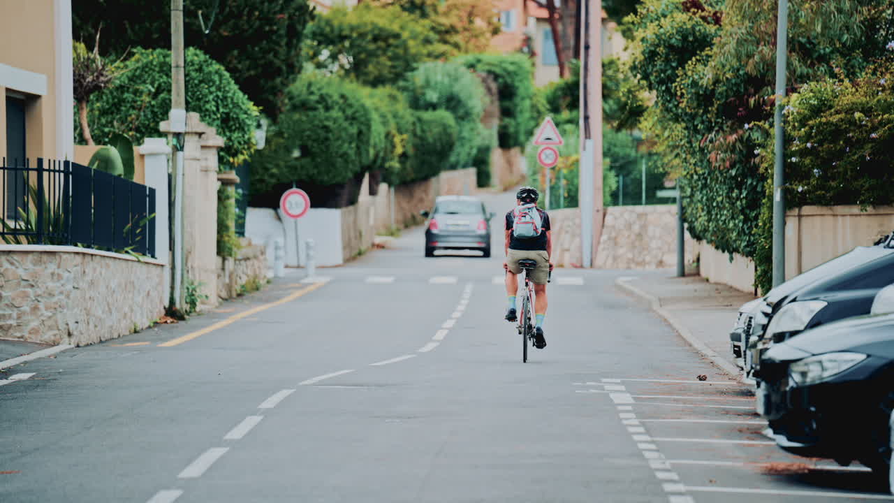 Rear view of a cyclist wearing a helmet and backpack riding along a calm suburban street lined with houses, trees and parked cars in soft daylight