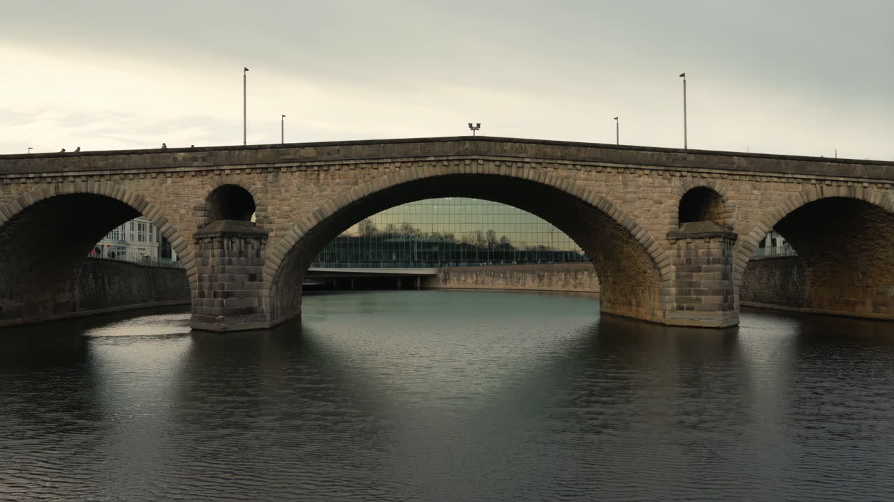 A historic stone arch bridge spans a calm river under an overcast sky, with a modern building visible through its central arch