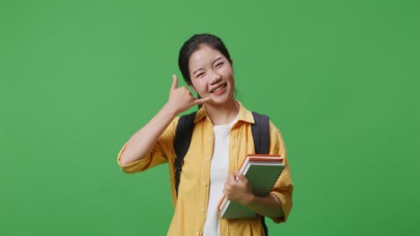 asian woman student with a backpack and some books smiling and showing call me gesture while standing in the green screen background studio