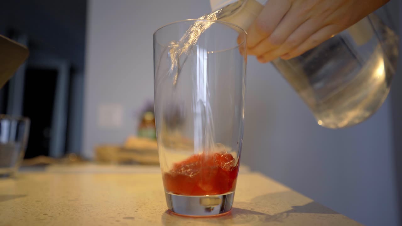 Pouring flavoured strawberry syrup into glass and filling with filtered water to created flavored water close-up selective-focus - Making syrup water in tall glass