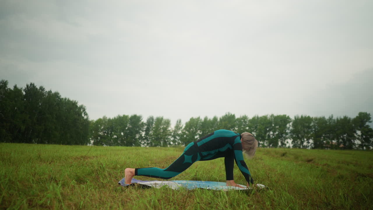 vista lateral de una mujer en traje verde y negro al aire libre en un vasto campo de hierba en una alfombra de yoga practicando una postura baja, bajo un cielo nublado con árboles alineados en el fondo