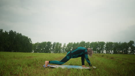 side view of woman in green and black suit outdoor in vast grassy field on yoga mat practicing low lunge pose, under cloudy sky with trees lined up in background