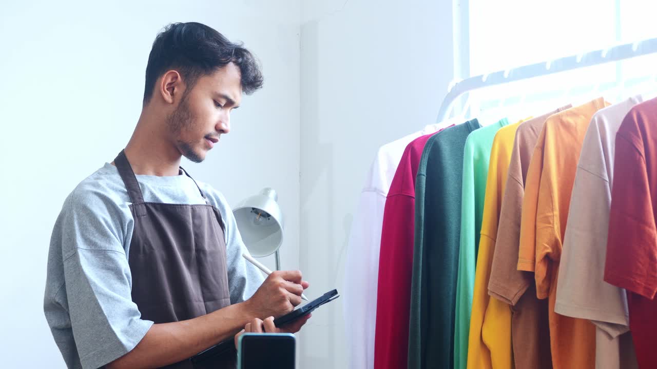 Young Asian Man In Apron Checklist While Inspects Clothing On Rack At Home Office