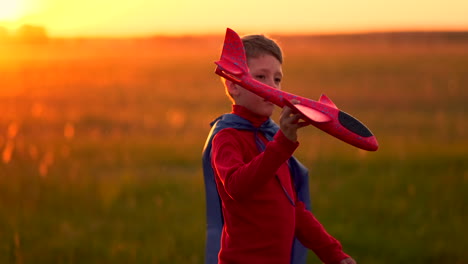 un niño feliz con una impermeable se imagina a sí mismo como un piloto de avión y corre con un juguete a través del campo pensando que es un piloto de aviones al atardecer.