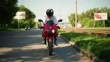 un motociclista con casco viaja rápido en una bicicleta eléctrica roja cerca de una carretera sin asfalto, los coches son visibles en el fondo borroso con su luz encendida, y también se ve un cartel en la distancia