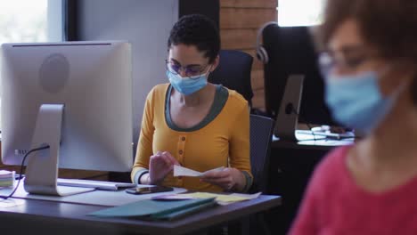 mujer caucásica con máscara de cara leyendo documentos y usando computadora en una oficina moderna
