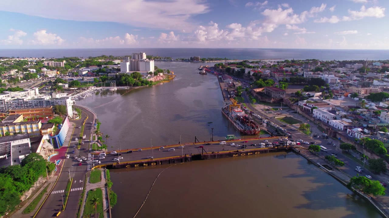 Toma aérea de dron en picado avanzando sobre el río Ozama y cruzando el Puente Flotante con tráfico vehicular hacia la histórica Zona Colonial y la salida al mar.