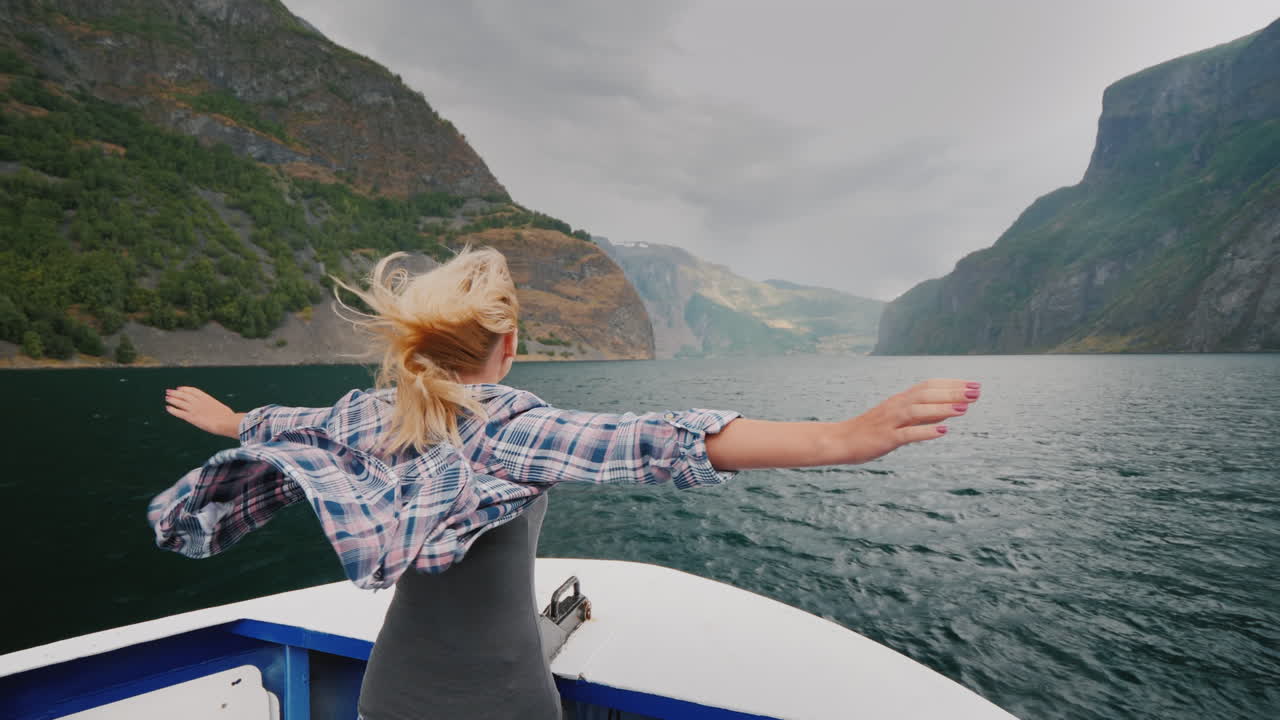 una mujer libre se para con las manos a los lados en la proa de un crucero que viaja por los fiordos de
