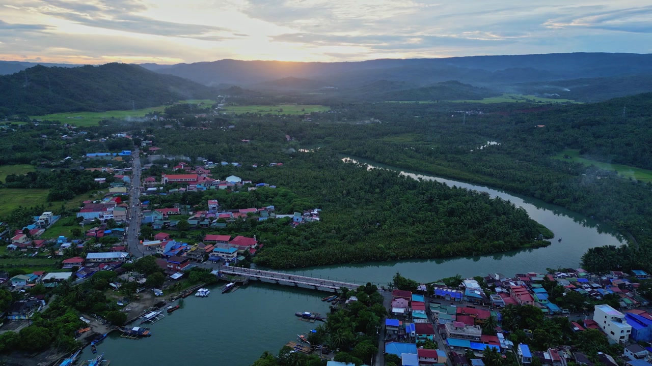 A slow forward drone shot glides over the provincial town, revealing colorful rooftops, winding river, and a warm sunset glowing behind the mountains of Mauban, Quezon Province Philippines