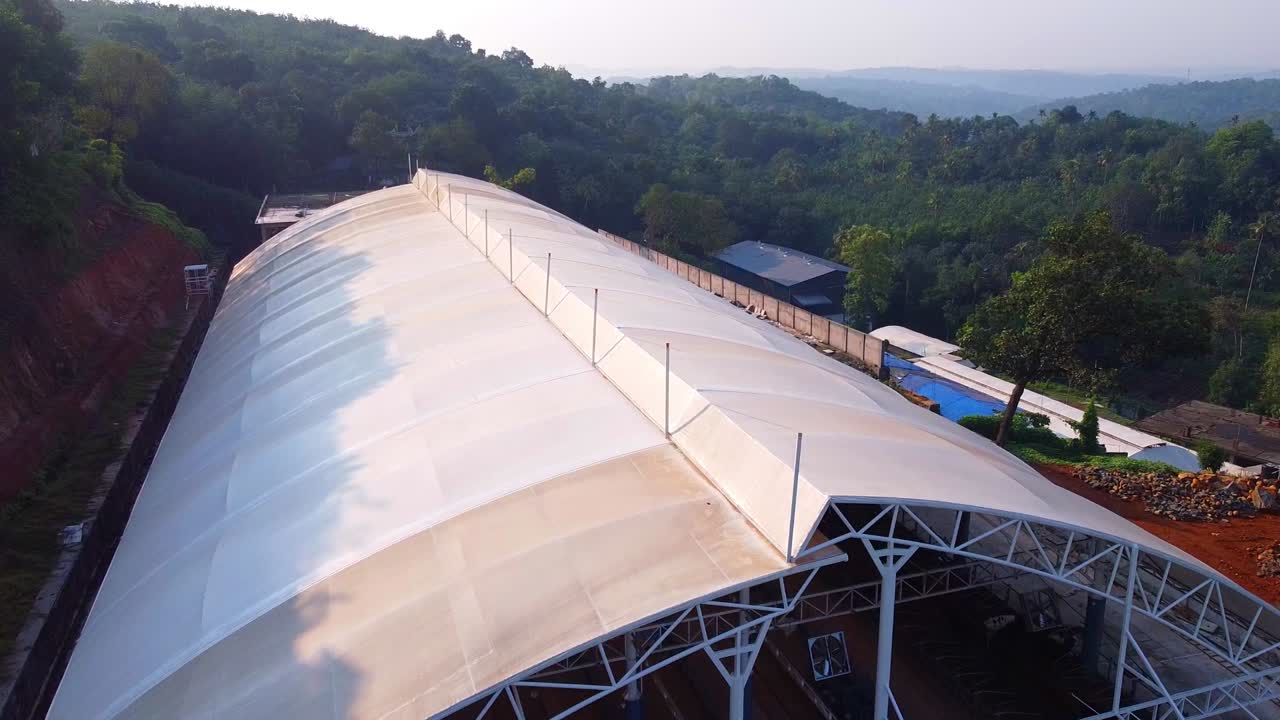 An aerial view of a large, modern greenhouse or industrial structure nestled in a lush, green hilly landscape, highlighting agricultural innovation and sustainable development