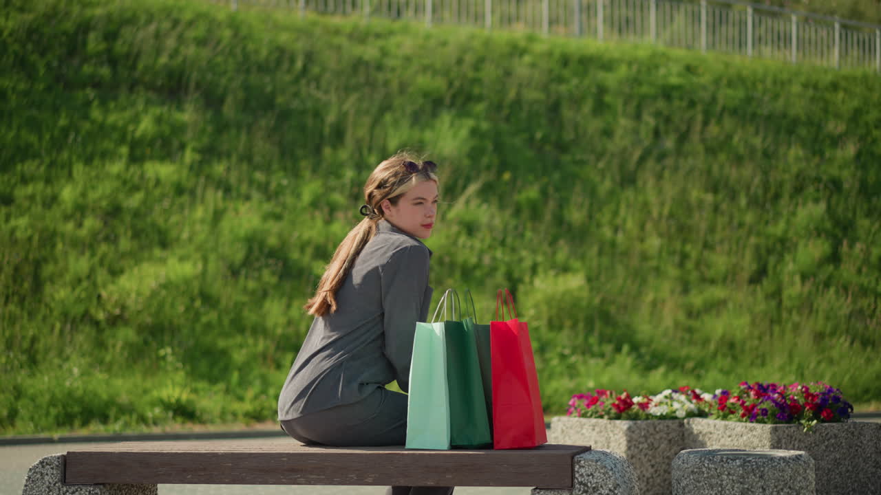 한 여인이 다채로운 쇼핑백을 들고 벤치에 다가와 앉아서 왼손을 벤치 위에 놓고 바람이 그녀의 머리카락과 초록색과 꽃을 흔들면서 자신을 조정합니다.