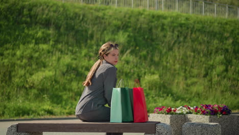 lady approaches bench carrying colorful shopping bags, sits down, relaxes her left hand on bench, and adjusts herself while wind flutters her hair, greenery and flowers in the background