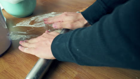 tortilla dough being flattened by two hands with a silver rolling pin on a flat wooden surface table during the daytime