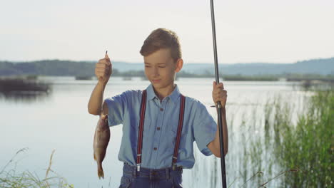 cute teenage boy posing for the camera with a fish on a rod while standing at the lake shore in the morning