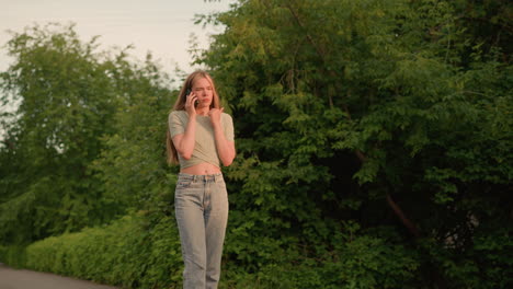 young woman in casual crop top holding phone to ear, walking on path surrounded by lush greenery with trees and shrubs, background includes distant car and building