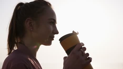 close up of a beautiful, dreaming young woman. girl is enjoying the moment. drinks from a sports mug. breathing. sun is shining on the background