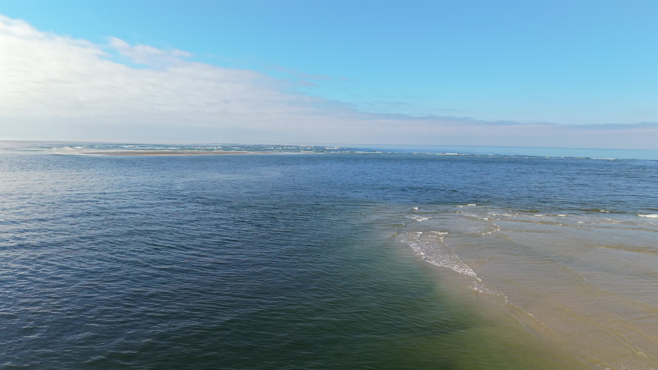Sandbanks rising from turquoise waters, forming narrow peninsula near Praia de Caieiras, Guaratuba, with urban skyline under cloudy blue atmosphere in Parana, Brazil