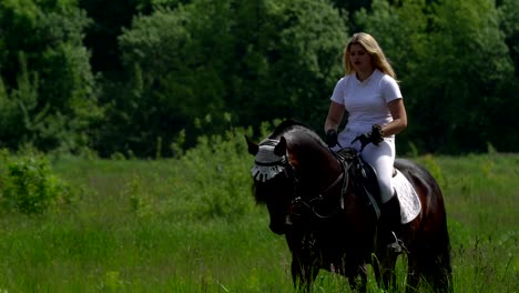 a beautiful girl in white hair and white clothes slowly rides on a black brown stallion at the racetrack. the girl warms up the horse before the performance.