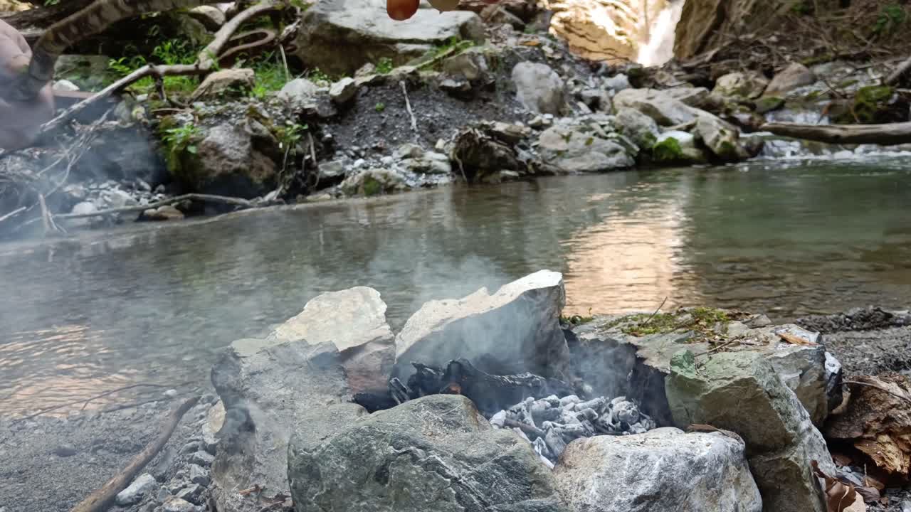 폭포를 배경으로 자연의 막대기에 세 개의 소시지를 요리하는 사람