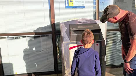 father teaching son how to vote with child casting letter in official ballot drop box for democratic government election