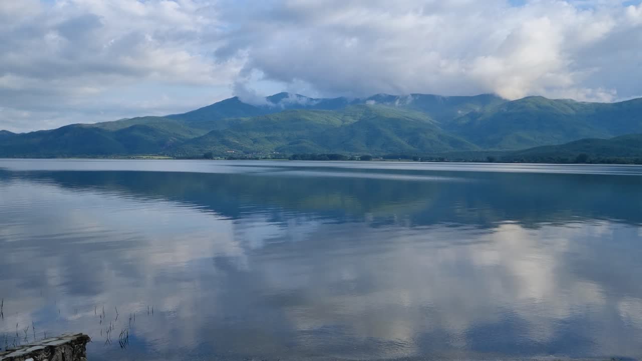 Scenic view of Kerkini Reservoir in northern Greece on a chilly spring day, with surrounding mountains and a cloudy sky reflecting in the still, clear waters