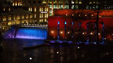 bridge and artificial waterfall in front of buildings. water illuminated in blue light.