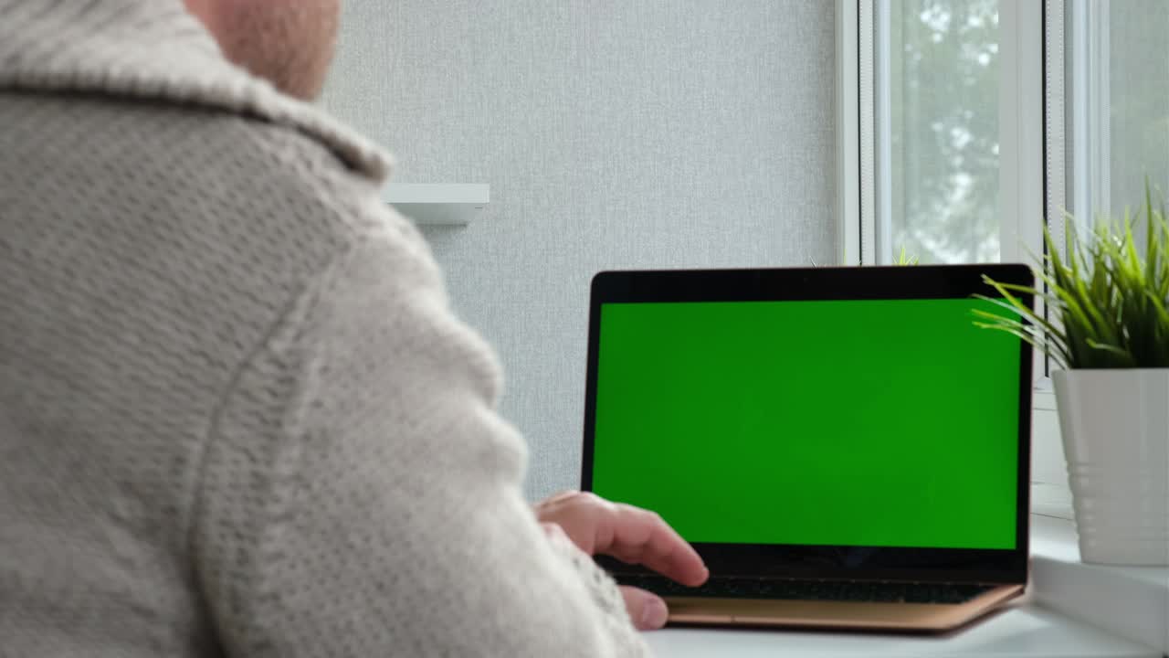 View from the back of man in warm sweater sitting at the table and typing on laptop with green chroma key screen. Laptop new technology concept. Freelancer working at home