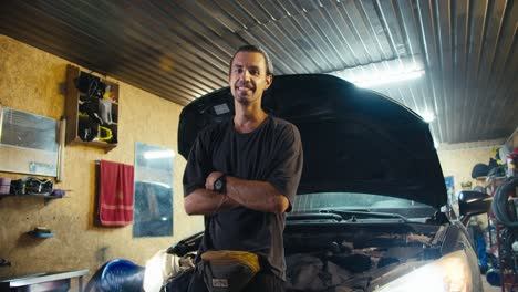 Portrait-of-a-confident-motorist-mechanic-who-puts-his-hands-on-his-chest-and-looks-at-the-camera-and-smiles-while-leaning-on-the-hood-of-his-car-in-a-workshop-garage-in-the-evening