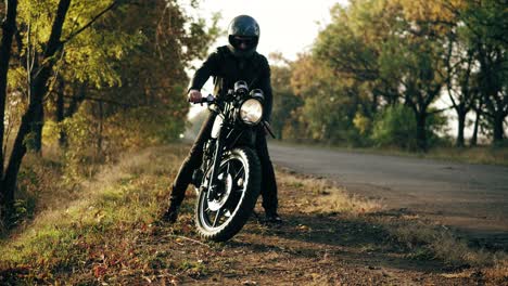 Unrecognizable-man-in-helmet-and-leather-jacket-coming-up-to-his-bike-and-starting-the-engine-while-standing-on-the-roadside-in-a-sunny-day-in-autumn