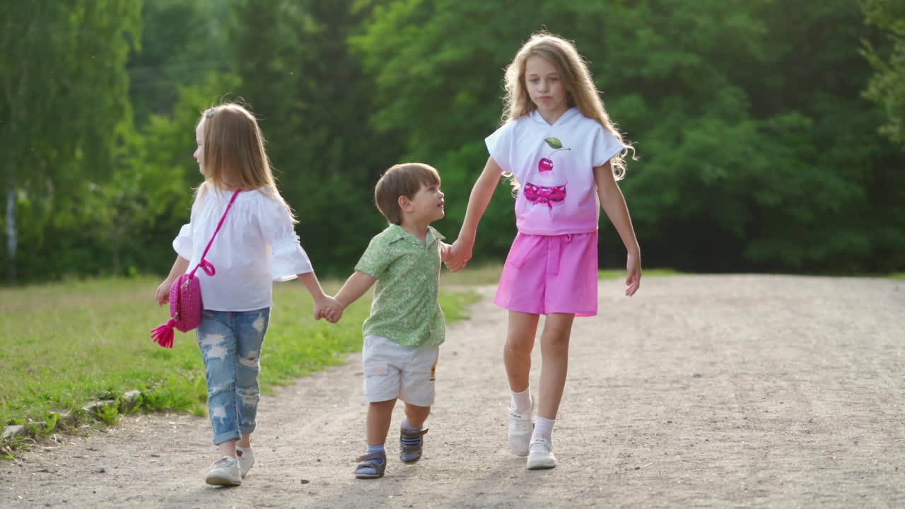 Two girls and one boy walk happily on the ground road in park. Sisters and brother have family day outdoor in summer. Concept video.