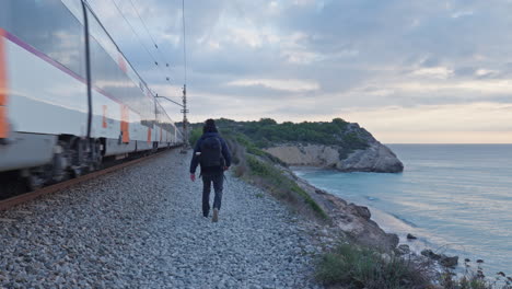 a man walks along train tracks as a passenger train rushes by nearby, while the calm sea washes down the cliff side