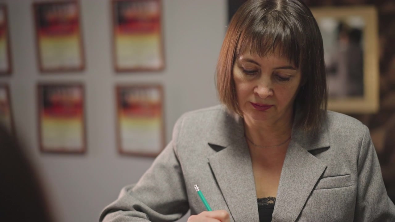 Amministratrice di scuola di musica che compila un modulo di iscrizione al banco della reception, una donna professionista e composta che firma documenti con una penna verde, certificati incorniciati alla parete, illuminazione calda dell'ufficio