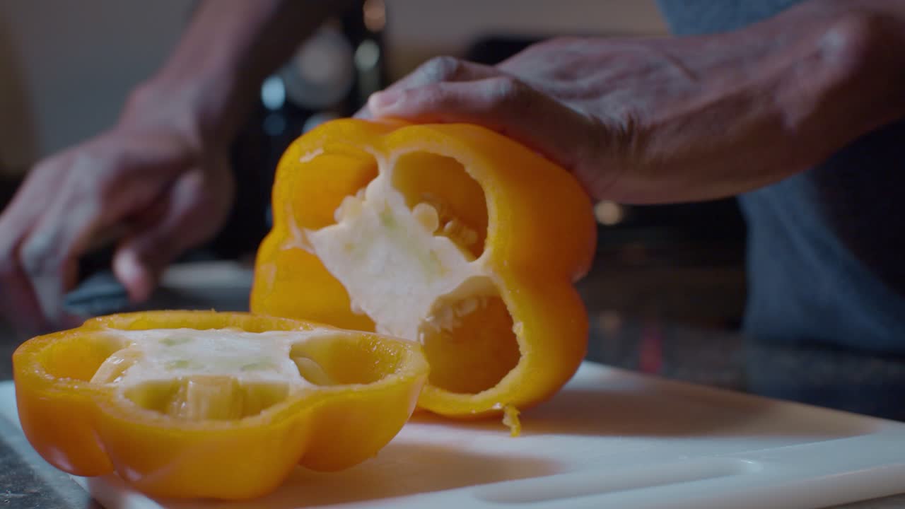 hombre negro cortando pimiento amarillo fresco en dos en una tabla de cortar blanca de plástico en la cocina