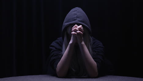 depressed person prays with tightly clenched hands wearing black hoody in a dark room