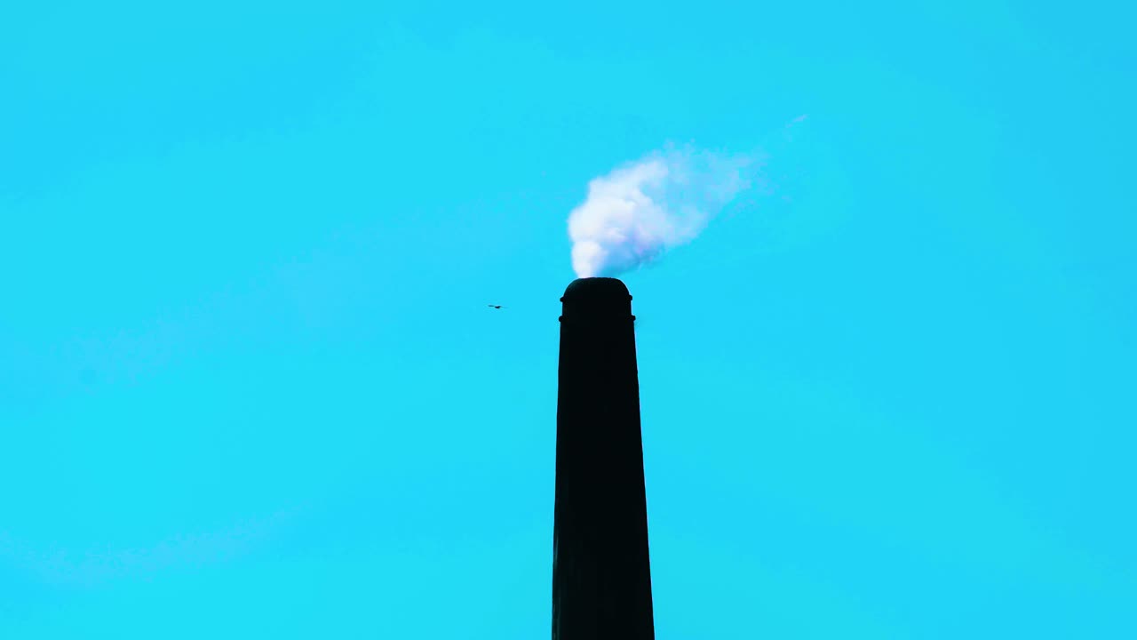 Static shot of a tall factory chimney releasing smoke at blue sky background