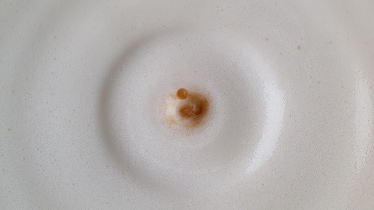 Goccia di caffè che gocciola nel latte schiumato bianco, creando schizzi e increspature, in macro e al rallentatore.