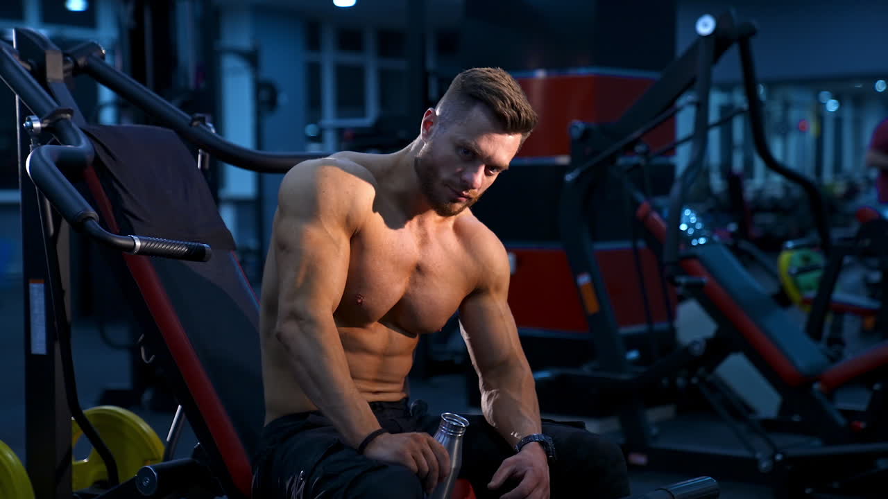 Handsome bodybuilder man drinks water in gym. Male athlete rests on bench. Slow motion.