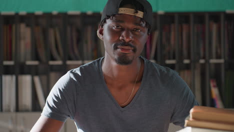 young man in cap sits at library table with stack of books, teal wall and organized shelves in background, lowering gaze before lifting head slowly, bathed in light and showing calm studious demeanor