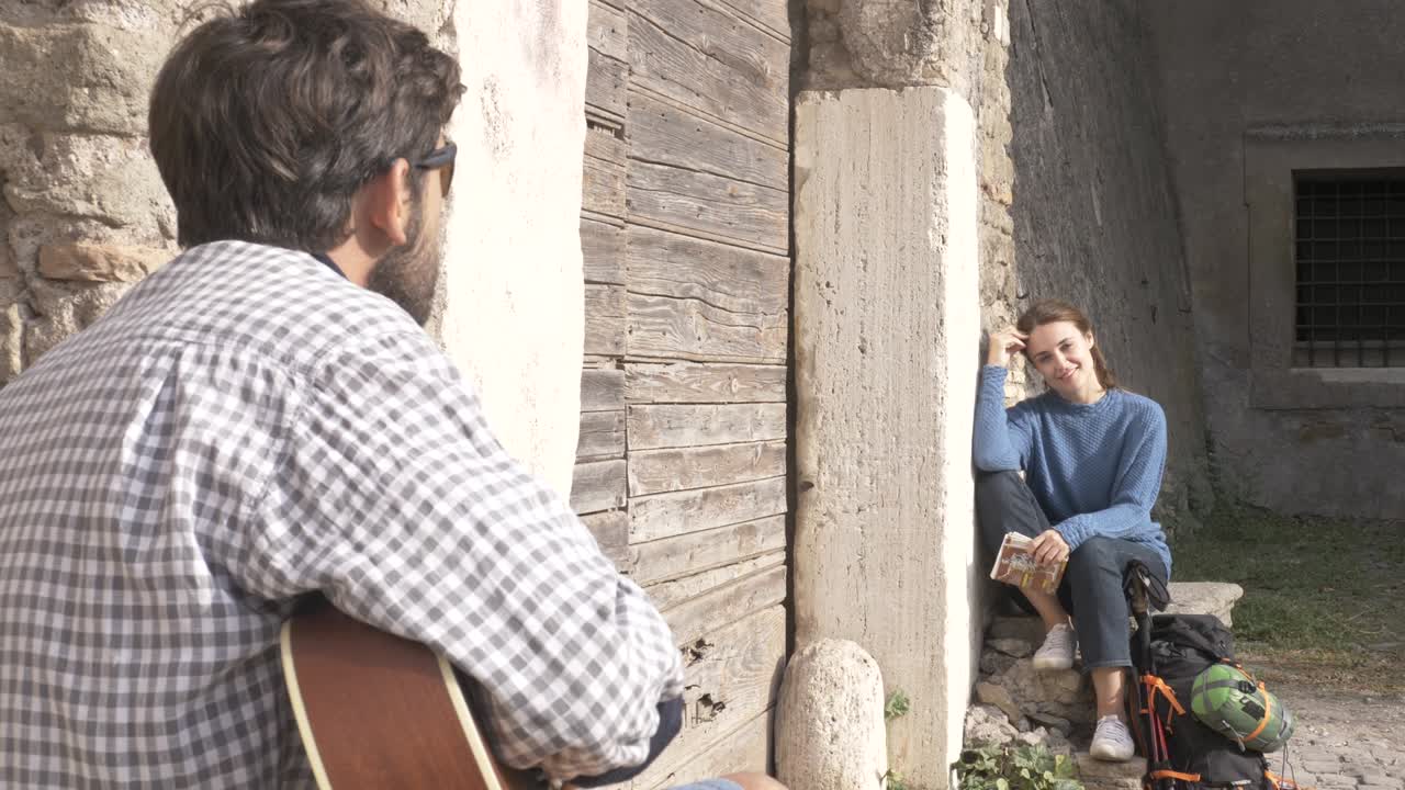 행복한 젊은 부부, 배 여행자, 기타를 연주하고 노래하는 관광객, 파르코 데글리 아쿠에도티의 중세 건물의 나무 문 근처에 앉아, 지도 가이드와 함께 해가 뜨는 로마의 파크 폐허, 느린 동작 스티디캠, 평면, 등급이 없습니다.