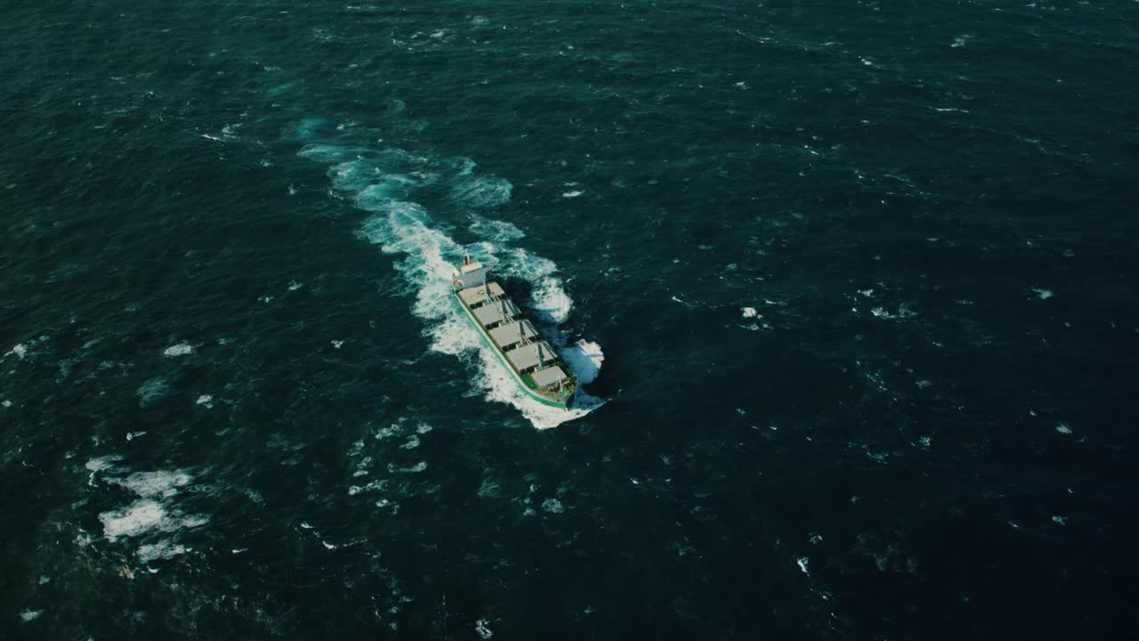 Large cargo ship sailing through rough ocean waves under cloudy sky, side view of bulk carrier with onboard cranes and rusted hull, maritime transport and global shipping concept