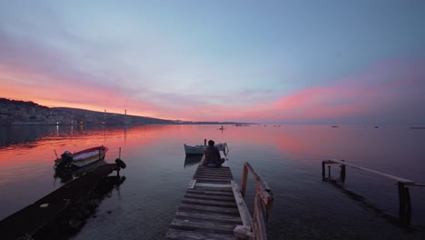 Hombre-Refugiado-Sentado-Con-Las-Piernas-Cruzadas-Medita-Observa-La-Increíble-Puesta-De-Sol-Desde-Una-Pequeña-Rampa-De-Madera-Para-Botes,-Lesbos,-Grecia,-Moria,-Lesbos