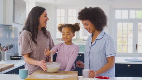familia con dos madres vestidas con pijama haciendo panqueques matutinos en la cocina en casa con la hija
