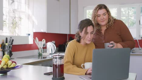 Feliz-Lesbiana-Caucásica-Tomando-Café-Y-Usando-Una-Computadora-Portátil-En-La-Cocina