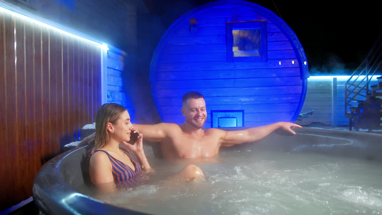 Relaxed Caucasian couple sit in the big Jacuzzi. Blonde woman speaks on the phone and man is smiling wide.