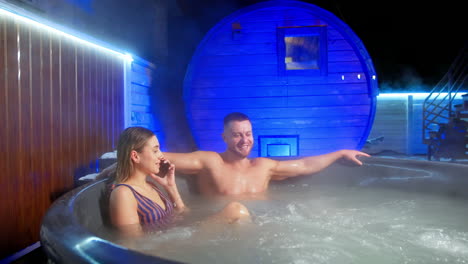 relaxed caucasian couple sit in the big jacuzzi. blonde woman speaks on the phone and man is smiling wide.