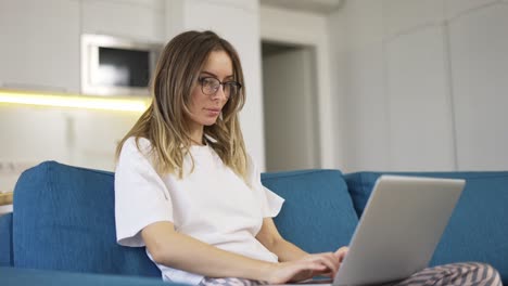 Blonde-woman-in-pajama-sit-on-the-couch-and-typing-on-silver-laptop,-low-angle-view