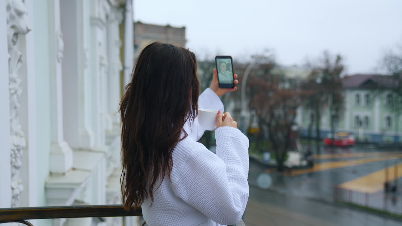 Young woman drinks coffee on a hotel balcony. Happy girl in a bathrobe doing selfie on the terrace on the urban background. Woman in white coat with a phone and a cup.